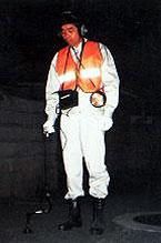 夜に反射板が付いたオレンジ色のベストを着た作業員がヘッドホンのようなものを耳にあて、黒い棒状の漏水探知機で調べている写真