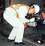 ヘルメットを被りオレンジ色のベストを着た作業員が腰を曲げた状態で音聴棒を使い調査している様子の写真